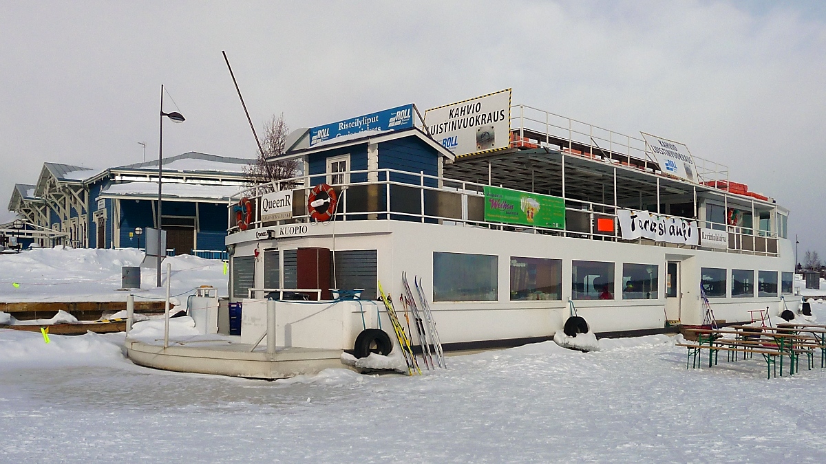 Auch festgefroren kann man ein Schiff noch nutzen. Das Ausflugsschiff  Queen R  dient als Cafeteria und Schlittschuh-Verleih (= Kahvio Luistinvuokraus) und bietet eine offene Terrasse mit Restaurant (= Terassi auki Ravintolalaiva) am Ufer des Kallavesi-Sees in Kuopio, Finnland, 8.3.13