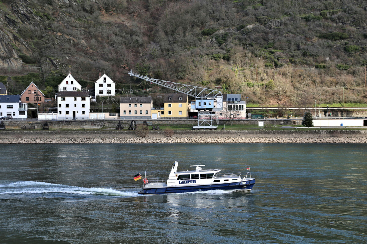 Auf dem Rhein stromaufwärts ist hier das Polizeiboot WSP 1 bei St. Goarshausen am 27.2.2023 um 14.20 Uhr unterwegs. Im Hintergrund ist ein alter Hafen Ladekran zu sehen.