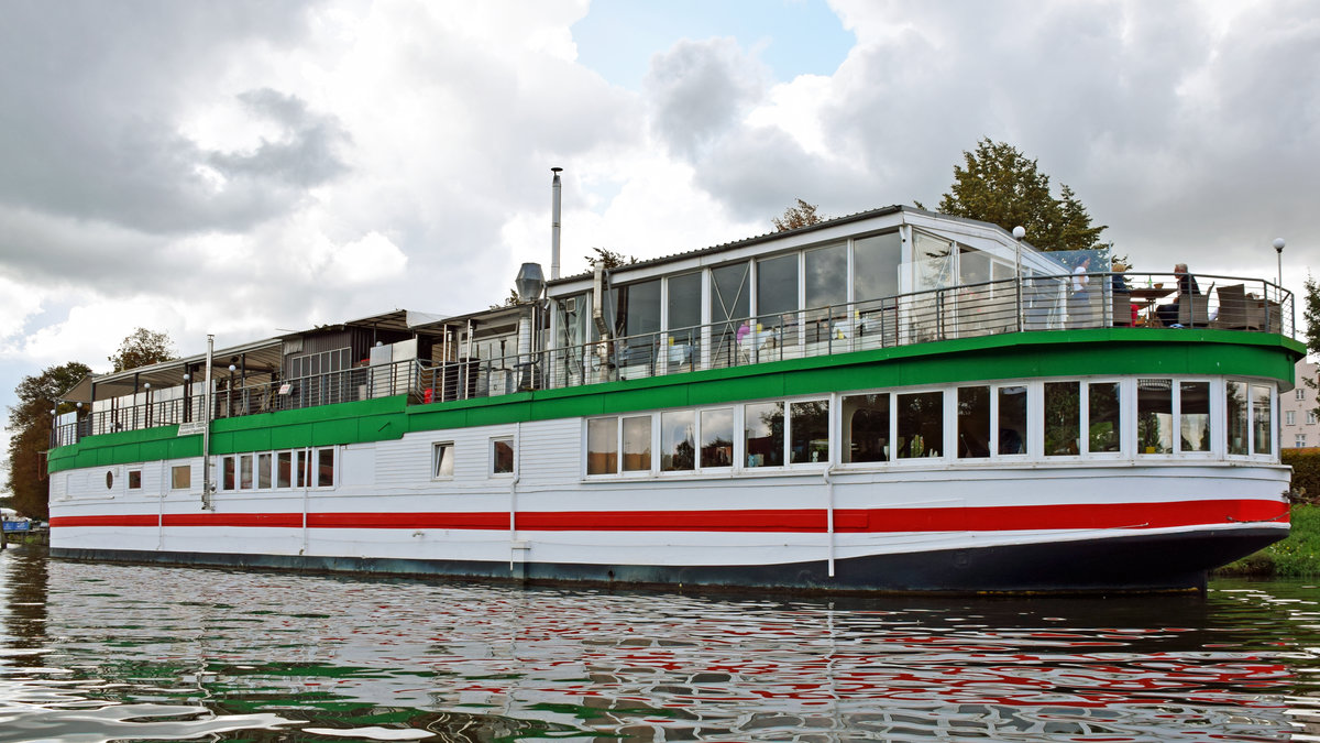 Das Betonschiff RIVERBOAT am 08.09.2019 in Lübeck. Es befindet sich seit den 1950er-Jahren in der Hansestadt und ist 55 Meter lang und 7,5 Meter breit. Es wurde 1942 von DYWIDAG (Dyckerhoff & Widmann) in Holland gebaut. Dieses Schiff bzw. dieser  Schwimmkörper , wie es korrekt heißen muss, denn es verfügte nie über einen eigenen Antrieb, wurde ausschließlich geschleppt und sollte schweres Kriegsgerät über den Kanal bringen. Nach Ende des Zweiten Weltkriegs diente es als Auffangstation für Flüchtlinge. 1956 wurde es unter der Lübecker Puppenbrücke vertäut und als Musikschiff (vor allem Jazz) eröffnet. 1978 wurde das Schiff auf die andere Seite des Holstentores, in den Holstenhafen, verbracht und als Diskothek wiedereröffnet. Als nach mehreren Anläufen auch hier der Erfolg langsam ausblieb, wurde das RIVERBOAT ein weiteres Mal verlegt;in den Klughafen (1994). Nach großen Umbau- und Sanierungsmaßnahmen öffnete im April 2007 das  neue  RIVERBOAT, jedoch nicht mehr als Diskothek, sondern als Veranstaltungszentrum. Das Restaurant  Seaside  bietet hier nunmehr  italienische Küche . 