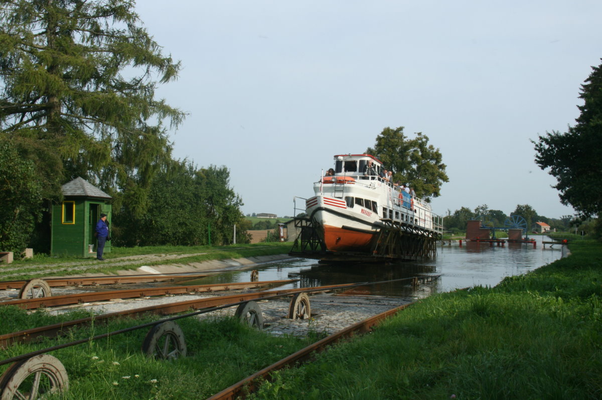 Die M/S  Marabut wird mit dem Trogwagen aus dem oberen Kanalteil gehoben und überquert in wenigen Augenblicken den Scheitelpunkt vom Rollberg Jelenie. Links im Bild sieht man den Wärter vom Rollberg vor seinem Wärterhäuschen. Die Rollbergwärter haben ihre Position am Scheitelpunkt der Rollberge. Von hier können sie sowohl den Schiffsverkehr im oberen und unteren Kanalbecken, als auch den Transport über den Rollberg beobachten. Mittels mechanischer Signaleinrichtung stehen sie in Verbindung mit dem Maschinisten im Maschinenhaus, was hinter dem Schiff zu erkennen ist. Im Hintergrund sieht man die Seilscheiben zum Umlenken des Zugseils vom Rollberg zur Fördermaschinen. Jelenie, 09.09.2017