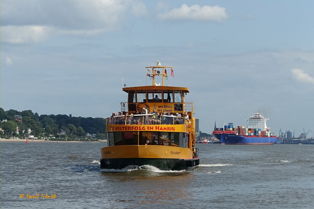 Hamburg am 21.8.2019: die HADAG-Fähre TOLLERORT steuert den Anleger Bubendeyufer an und im Hintergrund das auslaufende Containerschiff HERON HUNTER  /