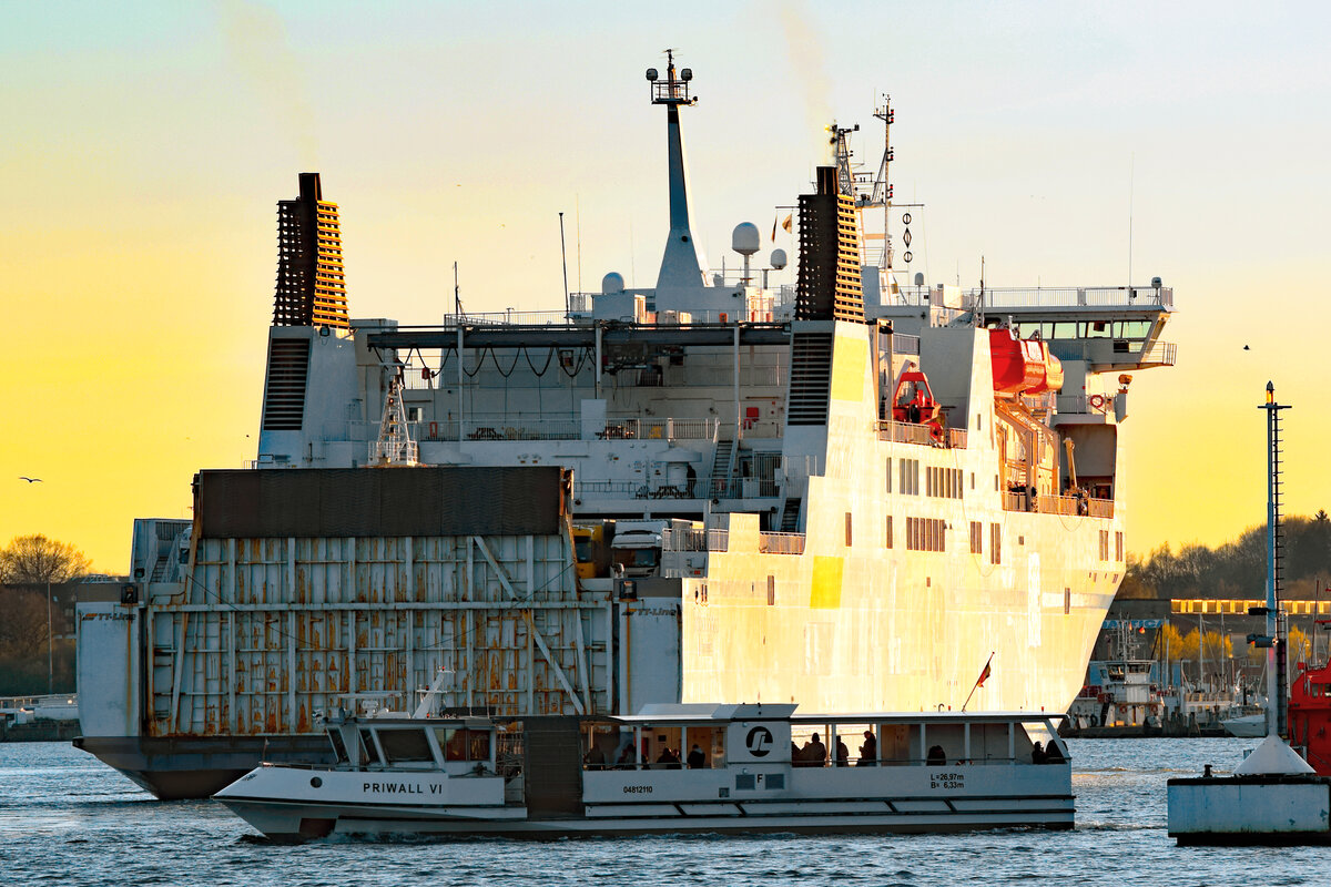 Priwall-Fähre PRIWALL VI quert am Abend des 16.04.2022 die Trave bei Lübeck-Travemünde. Die ebenfalls im Bild zu sehende TT-Line-Fähre ROBIN HOOD wird vom Licht der untergehenden Sonne illuminiert.