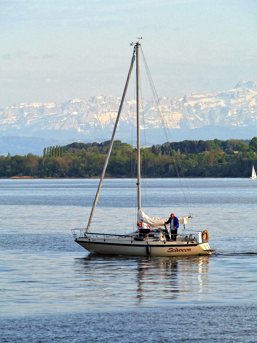 Segelboot  Scirocco  am Abend des 7. Mai 2016 mit eingeholtem Segel vor Überlingen. Während der Wald auf dem gegenüberliegenden Bodanrück wieder voll ergrünt ist, zeigen sich die Schweizer Alpen noch schneebedeckt. 