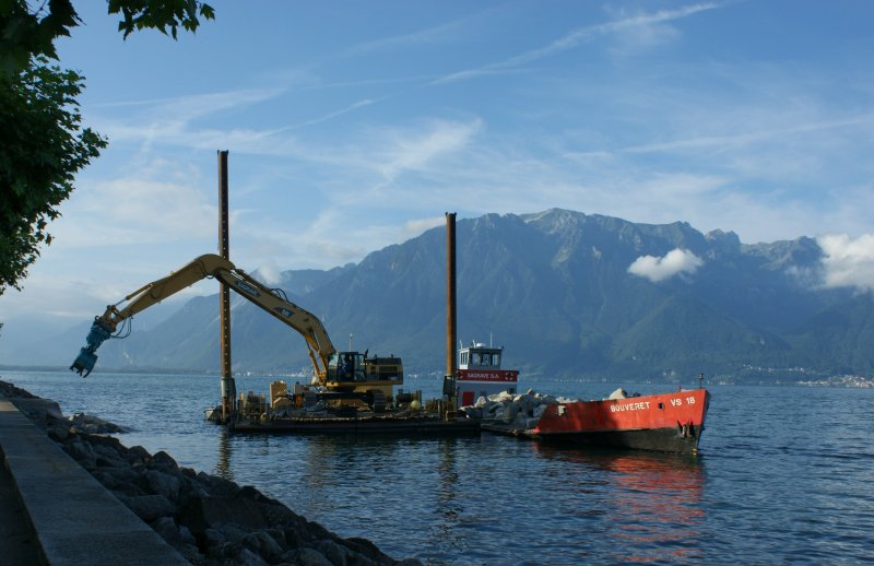 Der Kran auf dem Floss verteilt die Steine des Lastschiffes Bouvret an der Uferpromenade des Sees.
(August 2008)  