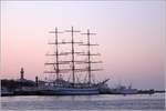 Das russische Segelschulschiff MIR (IMO 8501701) hat in Rostock-Warnemünde festgemacht. Vor dem abendlichen Himmel zeichnen sich Masten und Takelage ab. Links im Bild überragt der Leuchtturm Warnemünde die Häuser des Ortes. 10.11.2019